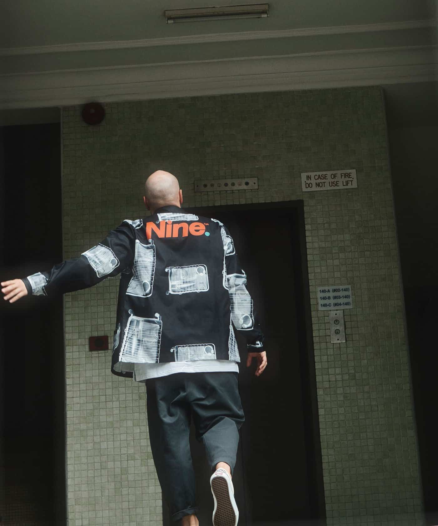 Man wearing a black streetwear jacket with white and red accents in an elevator lobby.